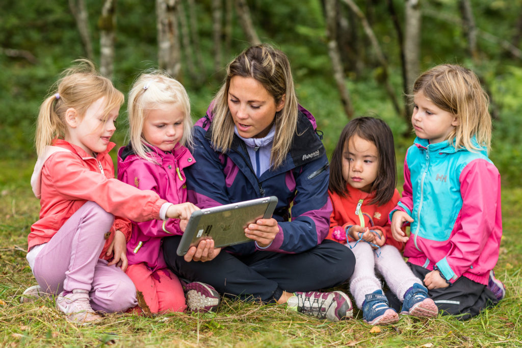 En voksen og fire barnehagebarn i naturen med iPad