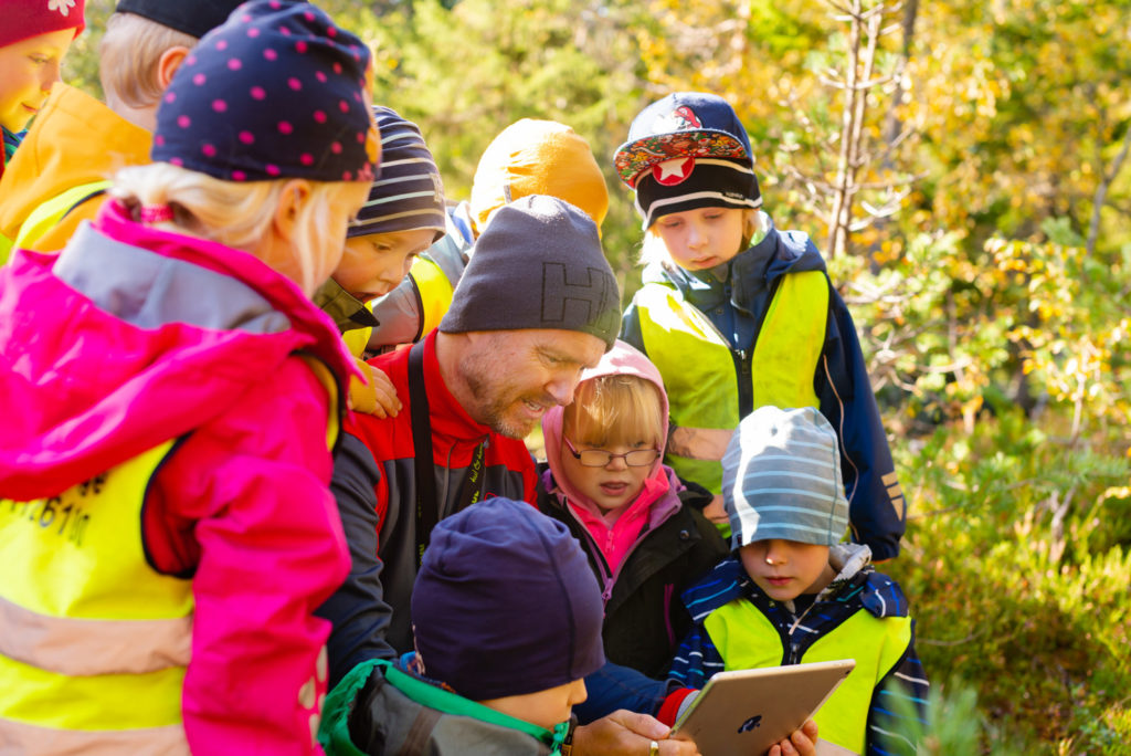 Barnehagebarn og en voksen ser på iPad i skogen